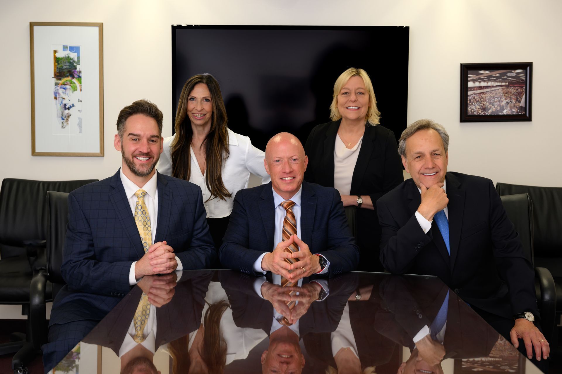 Five people in suits seated and standing around a dark table in an office.