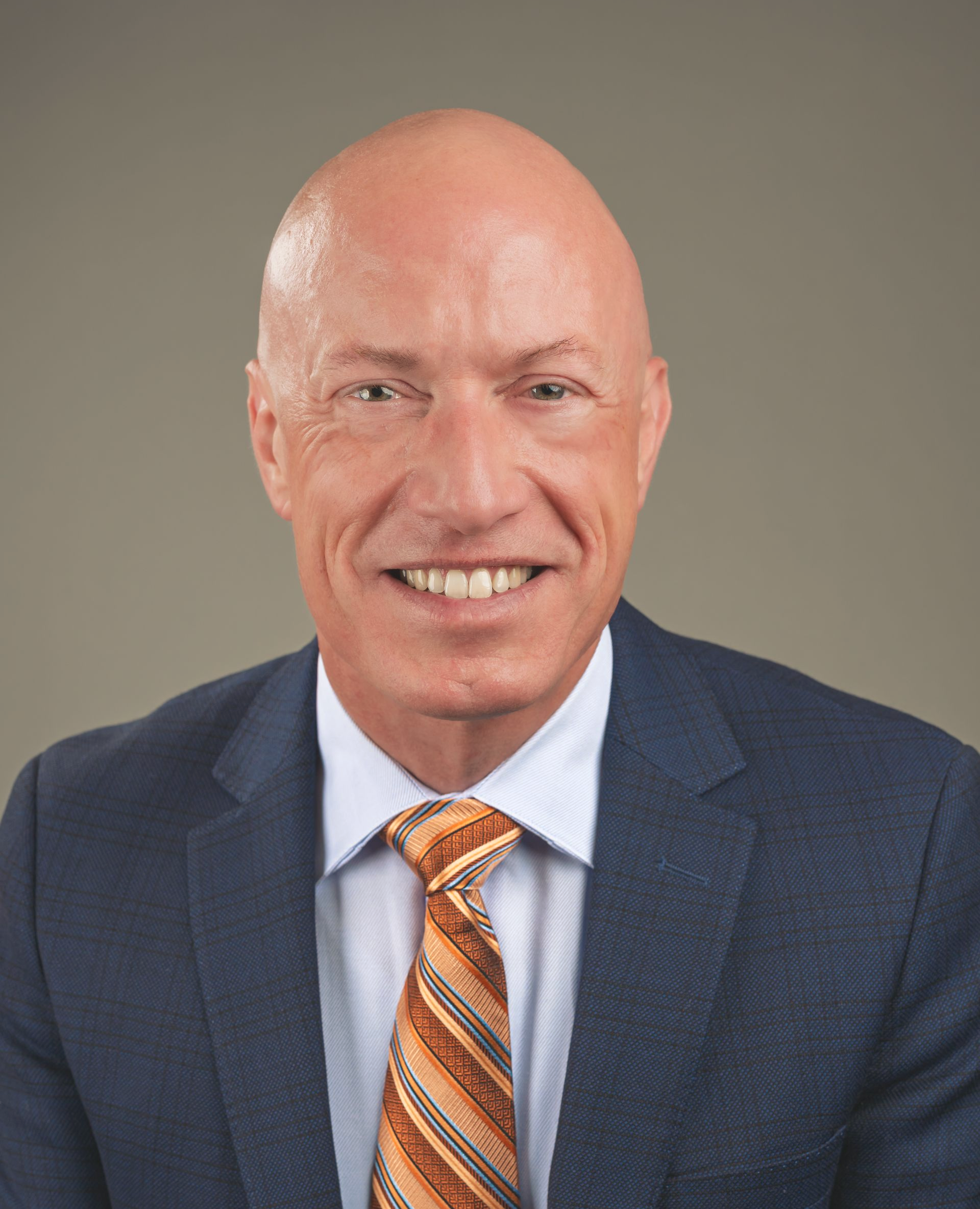 Bald man in a blue suit smiles at the camera, wearing an orange and brown tie.