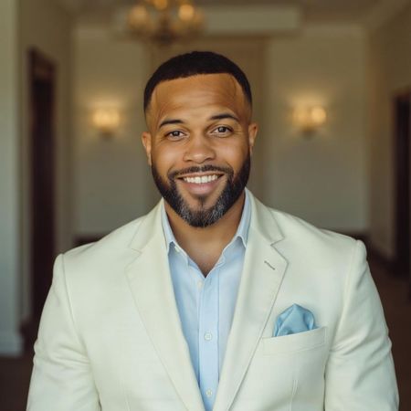 Smiling man in a light beige blazer and blue shirt, standing indoors with a softly blurred hallway background