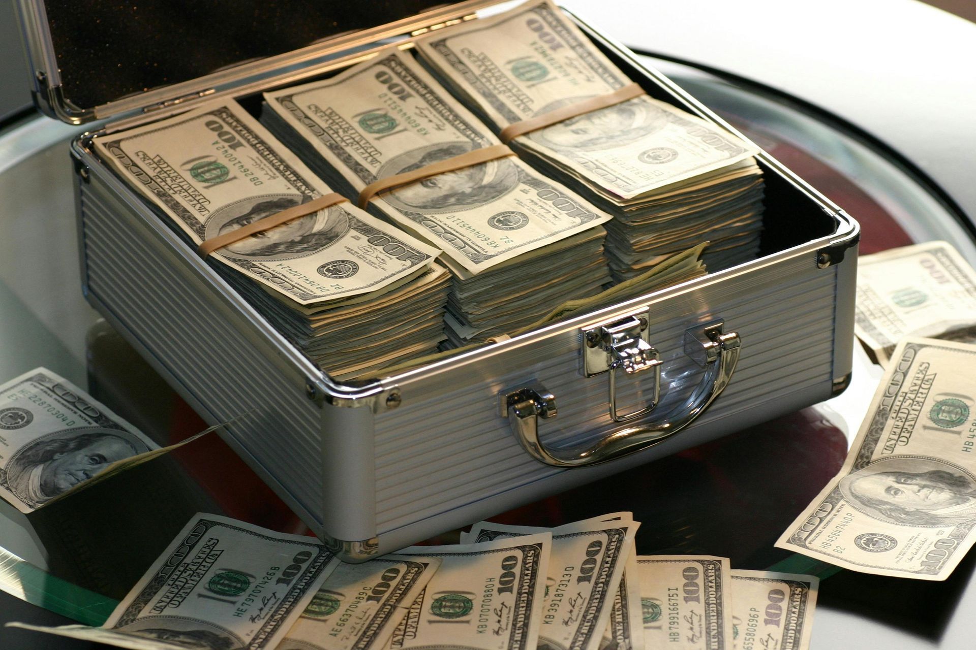 Silver briefcase overflowing with stacks of US $100 bills, with some scattered on the table.