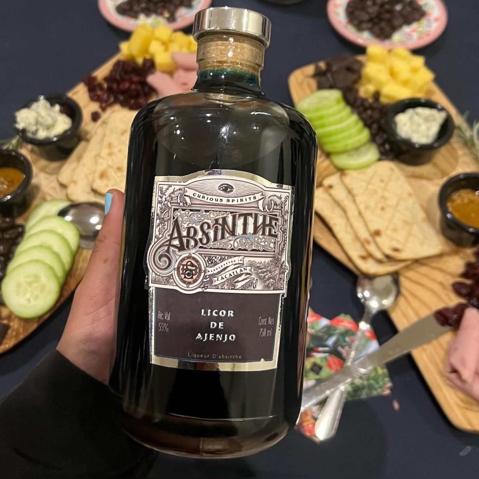 Una persona sostiene una botella de absenta frente a una mesa llena de comida.