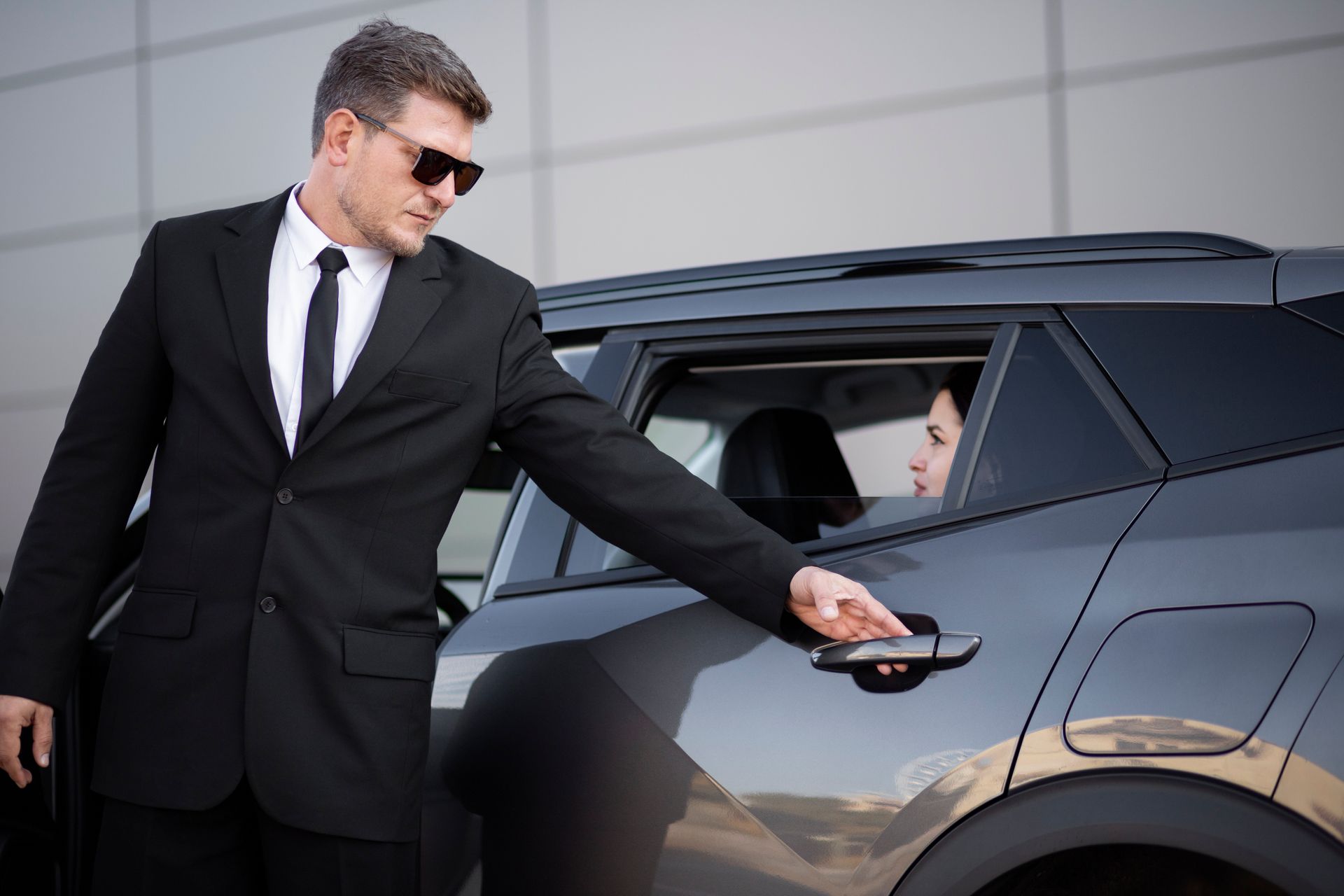 Un homme en costume noir ouvre la portière d'un SUV noir tandis qu'un passager est assis à l'intérieur.