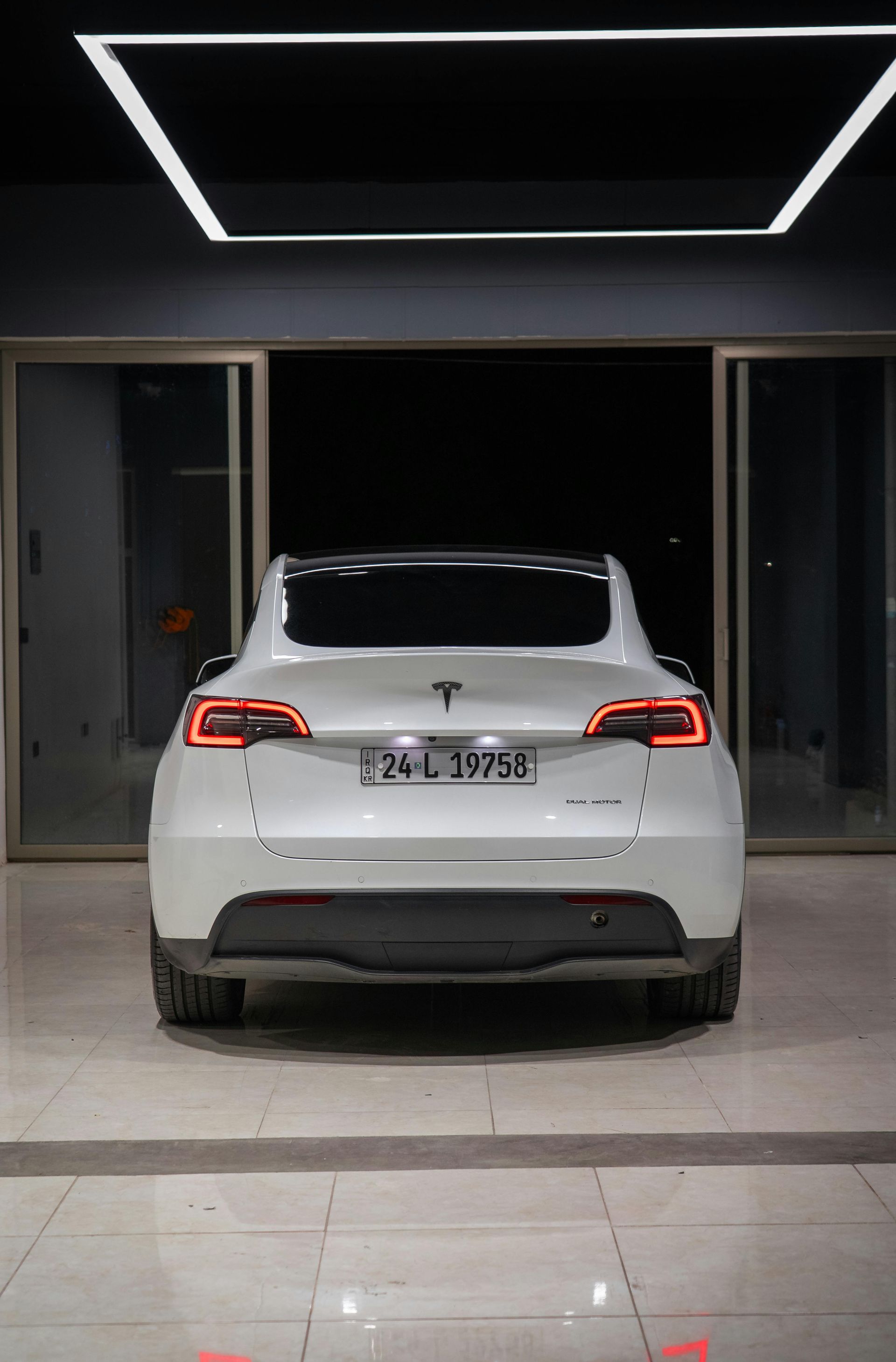 Une berline Tesla blanche est garée de nuit devant l'entrée d'un immeuble moderne (vue arrière).