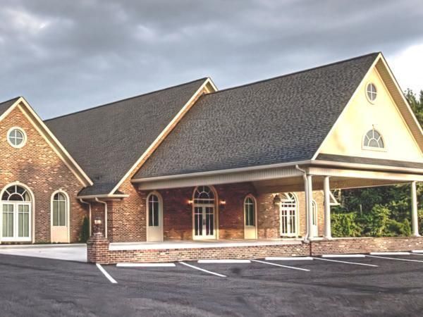 Brick church building with arched windows, white columns, and a dark roof beside an empty parking lot