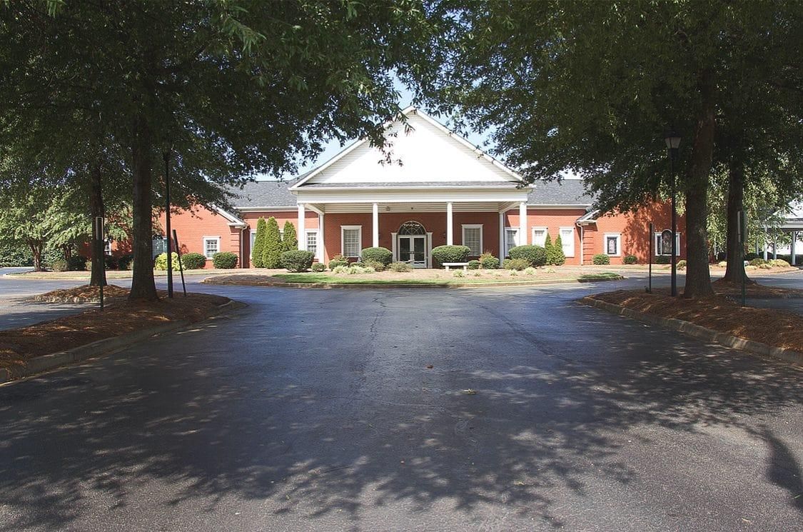 Brick building with white columns and red doors, framed by trees and a paved drive.