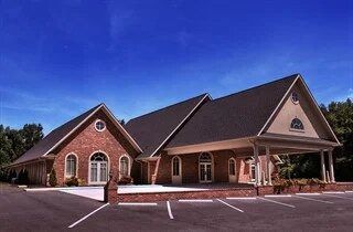 Brick church building with white-trimmed windows and a dark roof in a parking lot under a blue sky