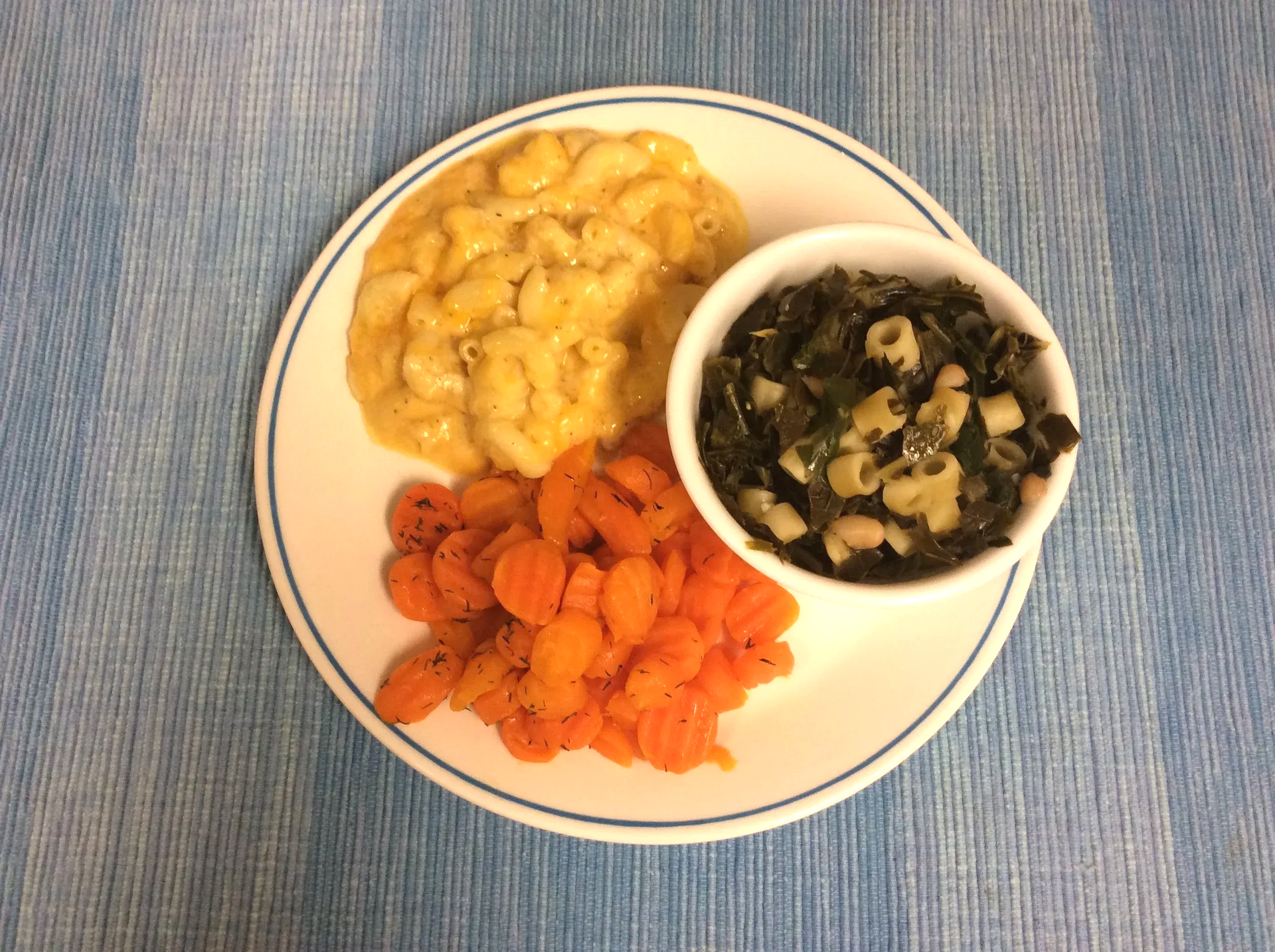Plate with macaroni and cheese, roasted carrots, and a bowl of leafy greens on a blue cloth.