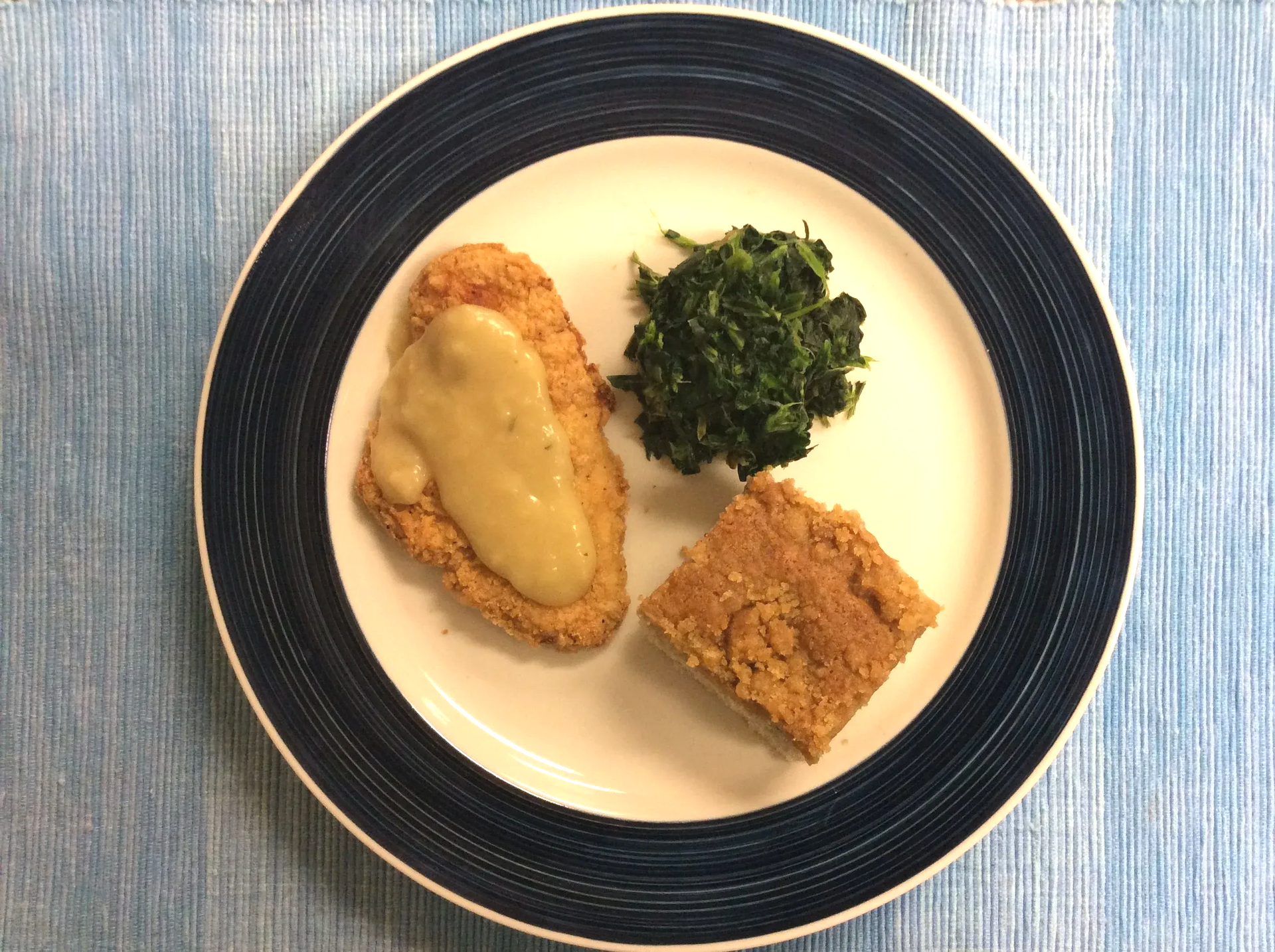 Three-piece meal on a white plate with dark rim: breaded cutlet, greens, and a square pastry