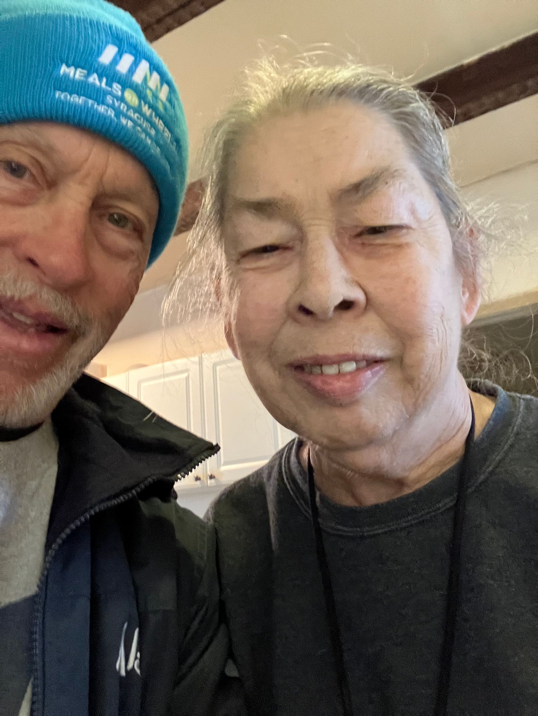 Two people smiling closely for a selfie indoors; one wears a blue beanie.