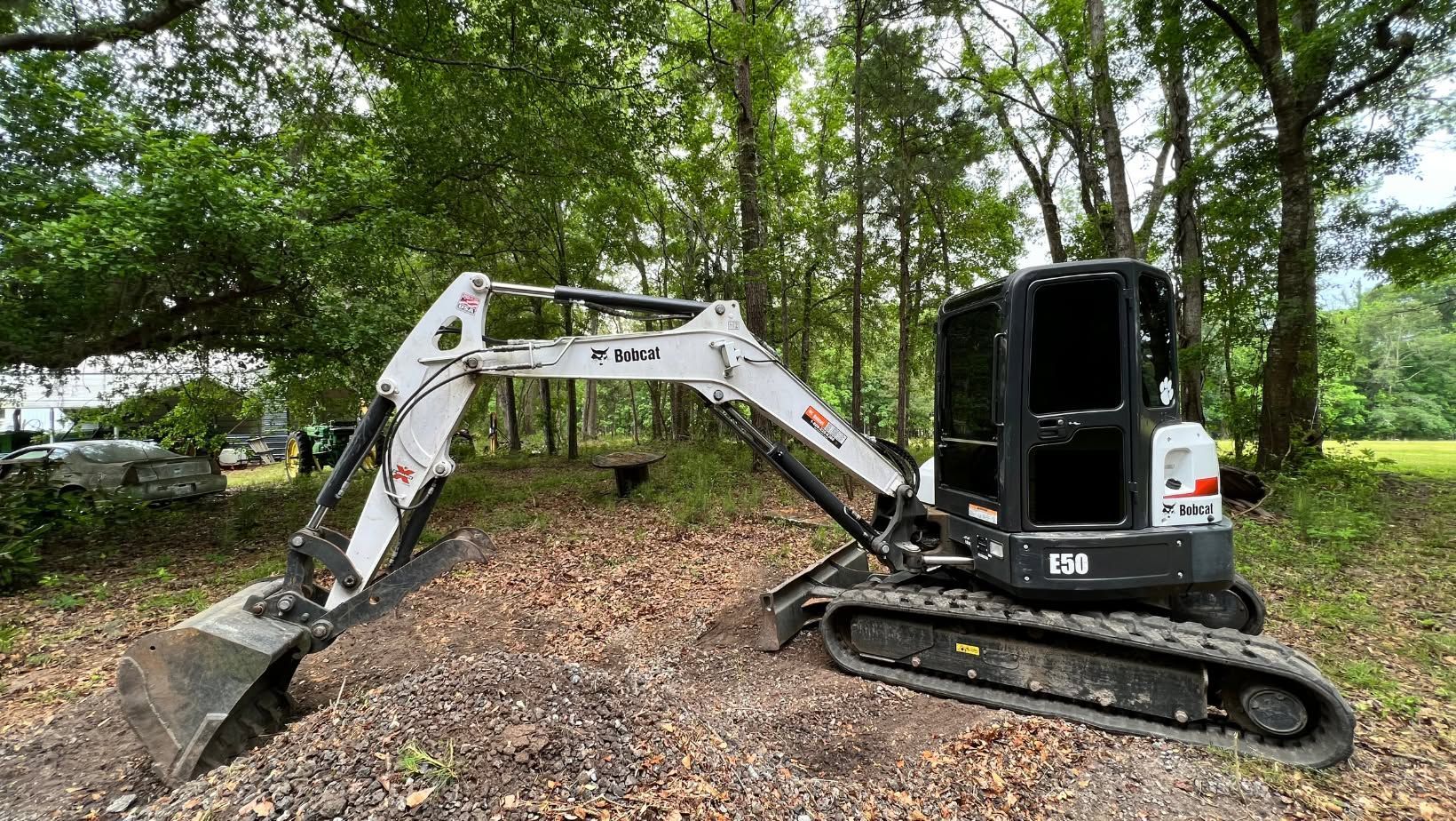 Small excavator with extended arm on a leaf-covered path in a wooded area