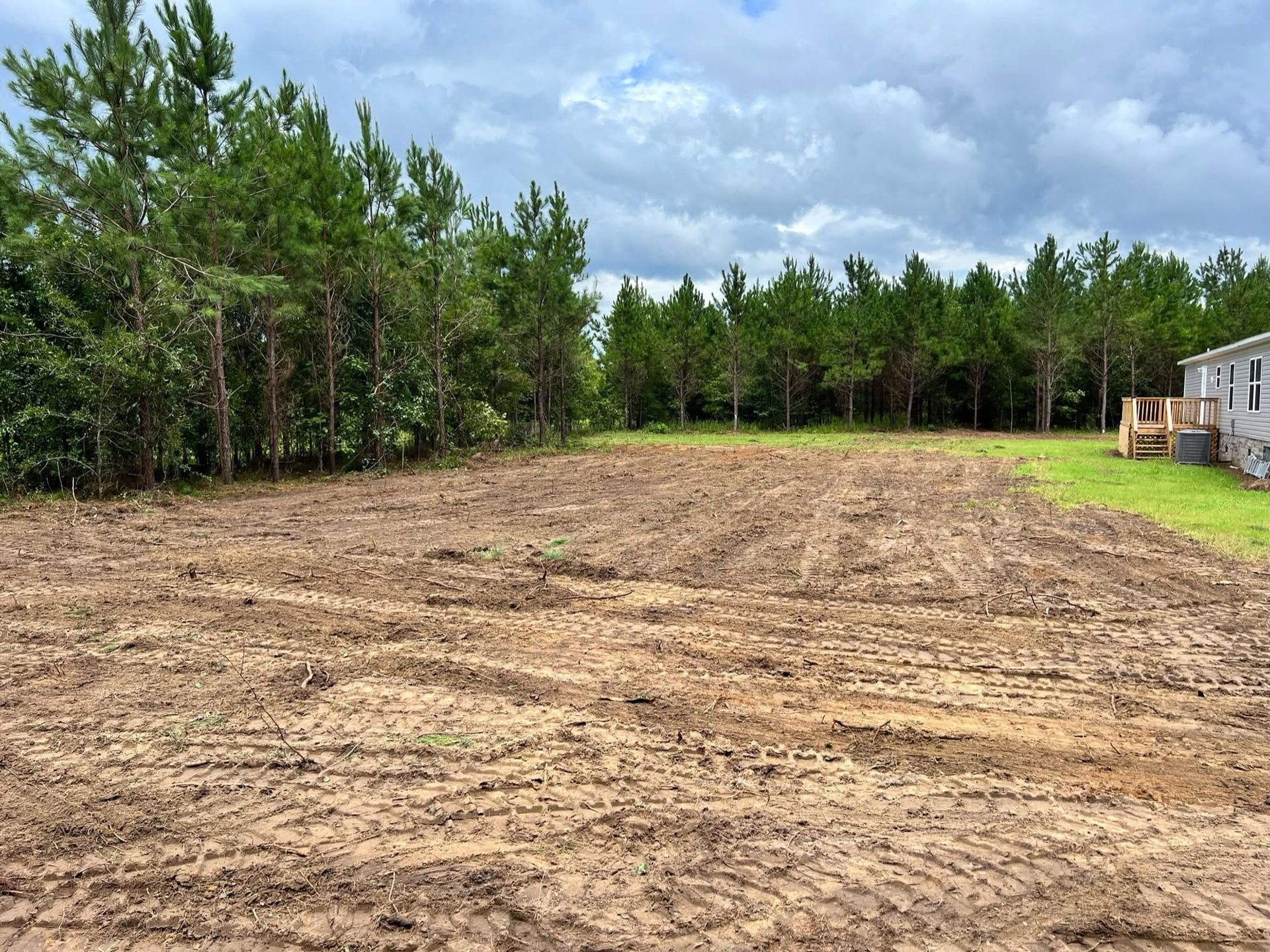 Cleared dirt lot beside a house, bordered by trees under a cloudy sky