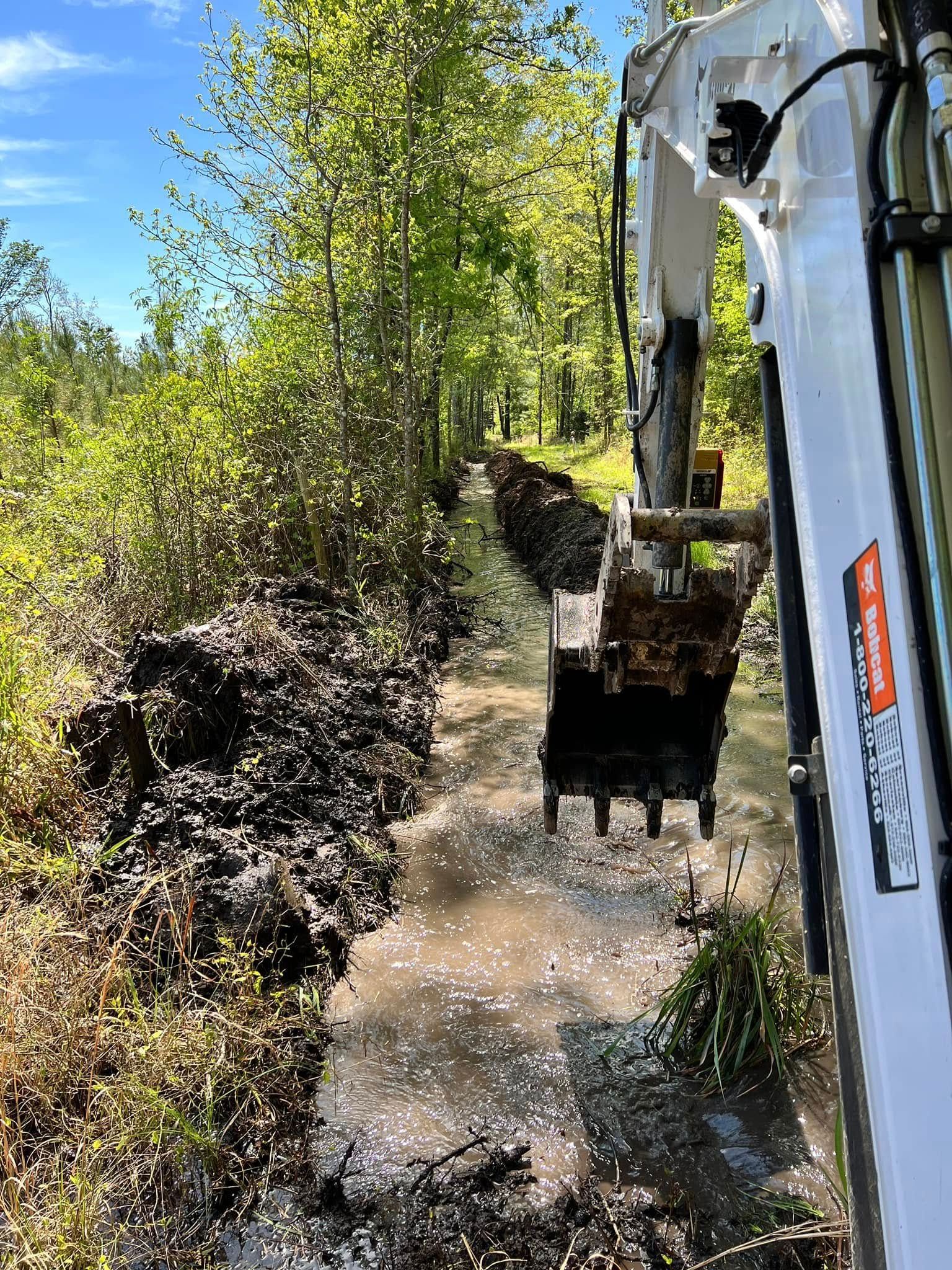 Backhoe loading muddy gravel beside a wooded trail on a sunny day