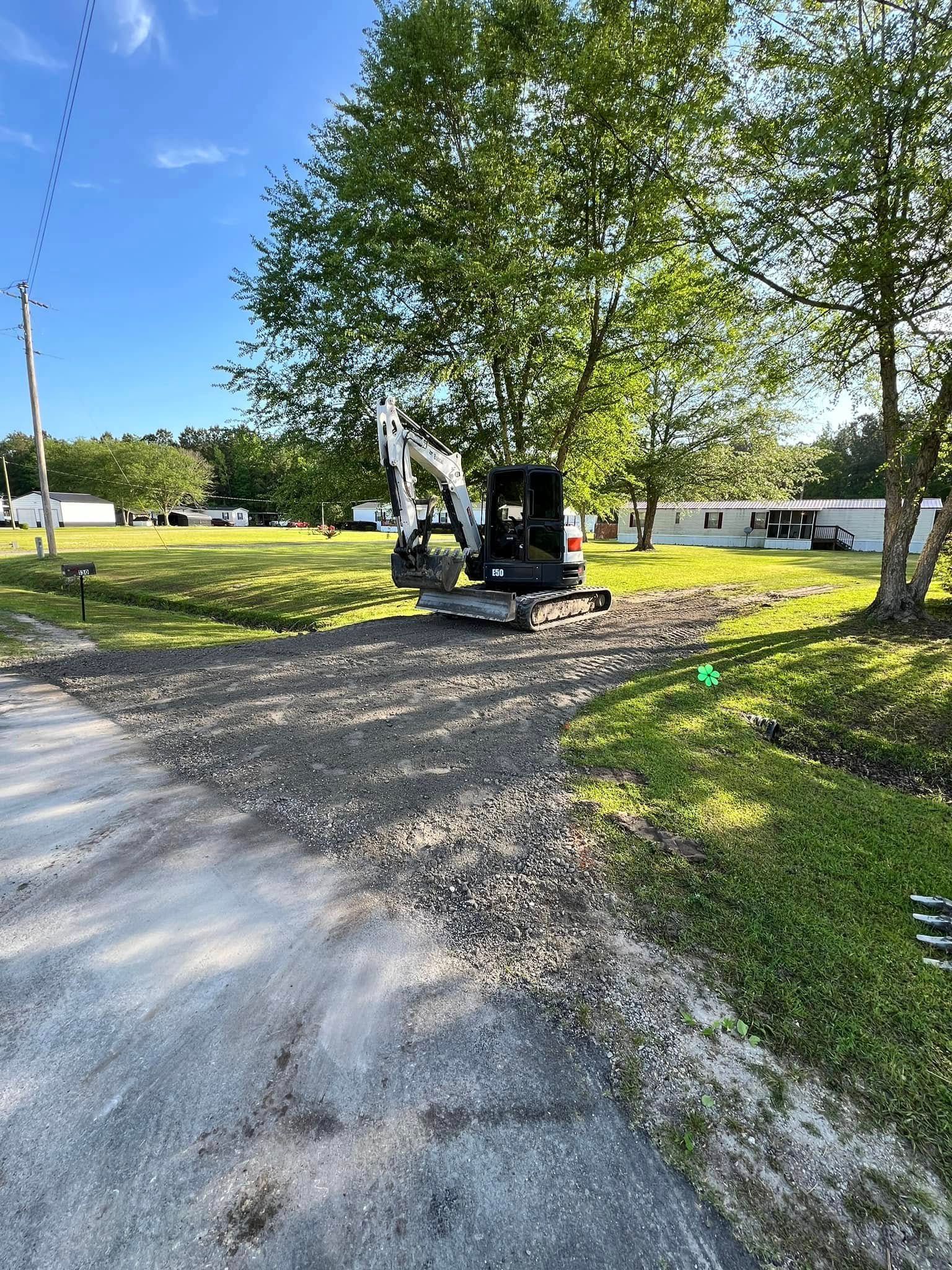 Excavator grading a gravel driveway beside a large tree on a sunny day