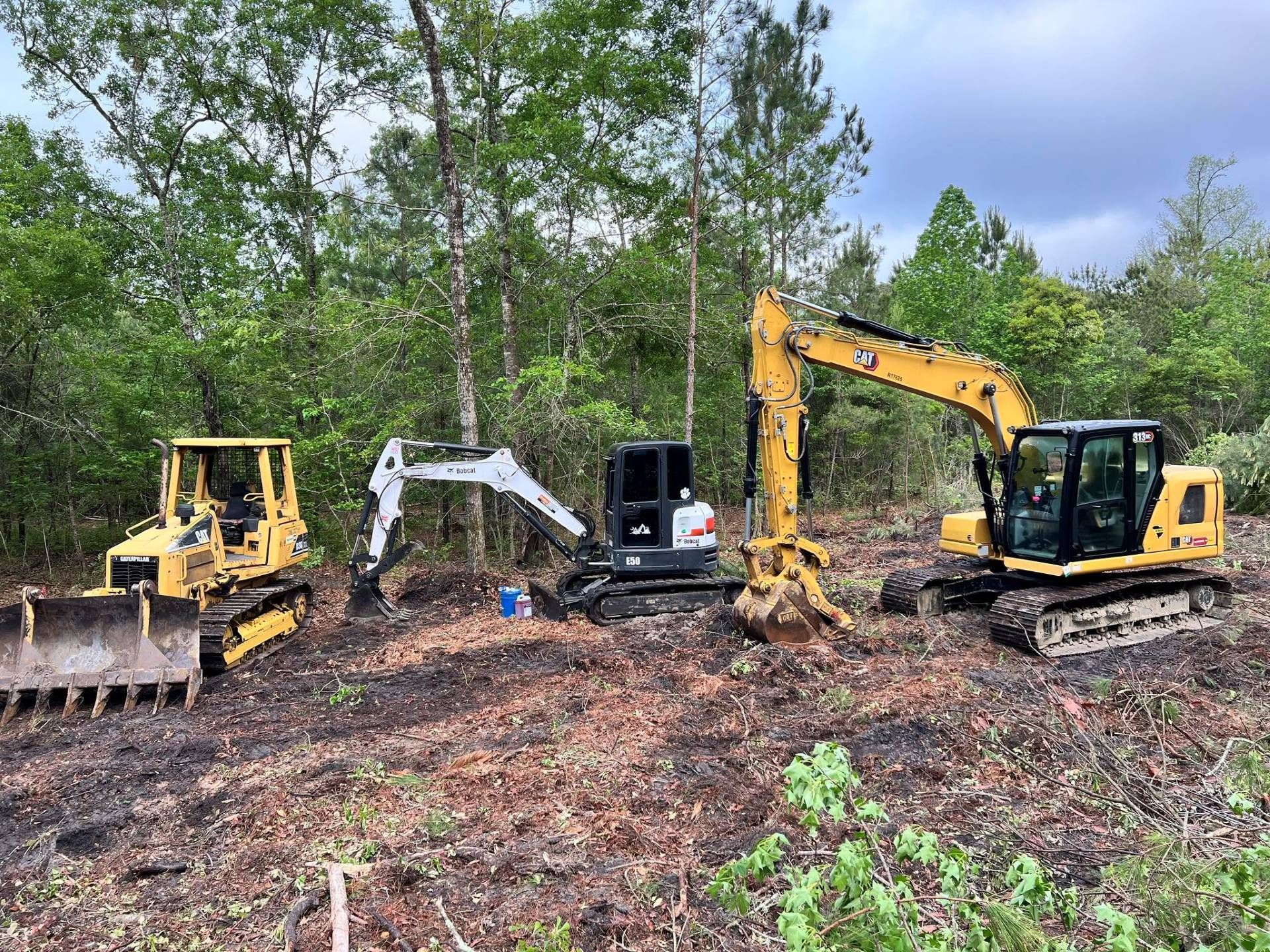 Three excavators clearing a muddy wooded site, including a yellow excavator and a white mini excavator.