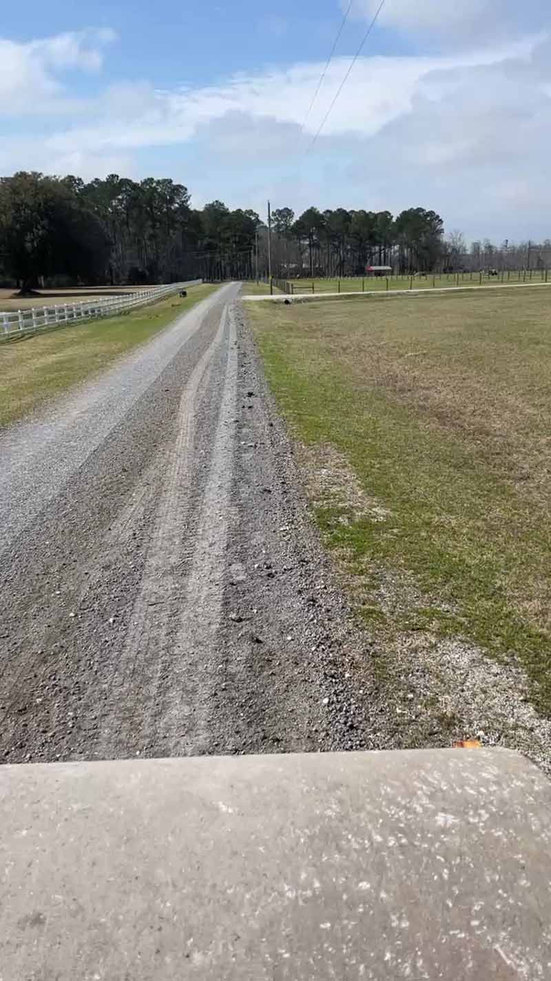 Gravel road through a grassy field with trees in the distance under a partly cloudy sky