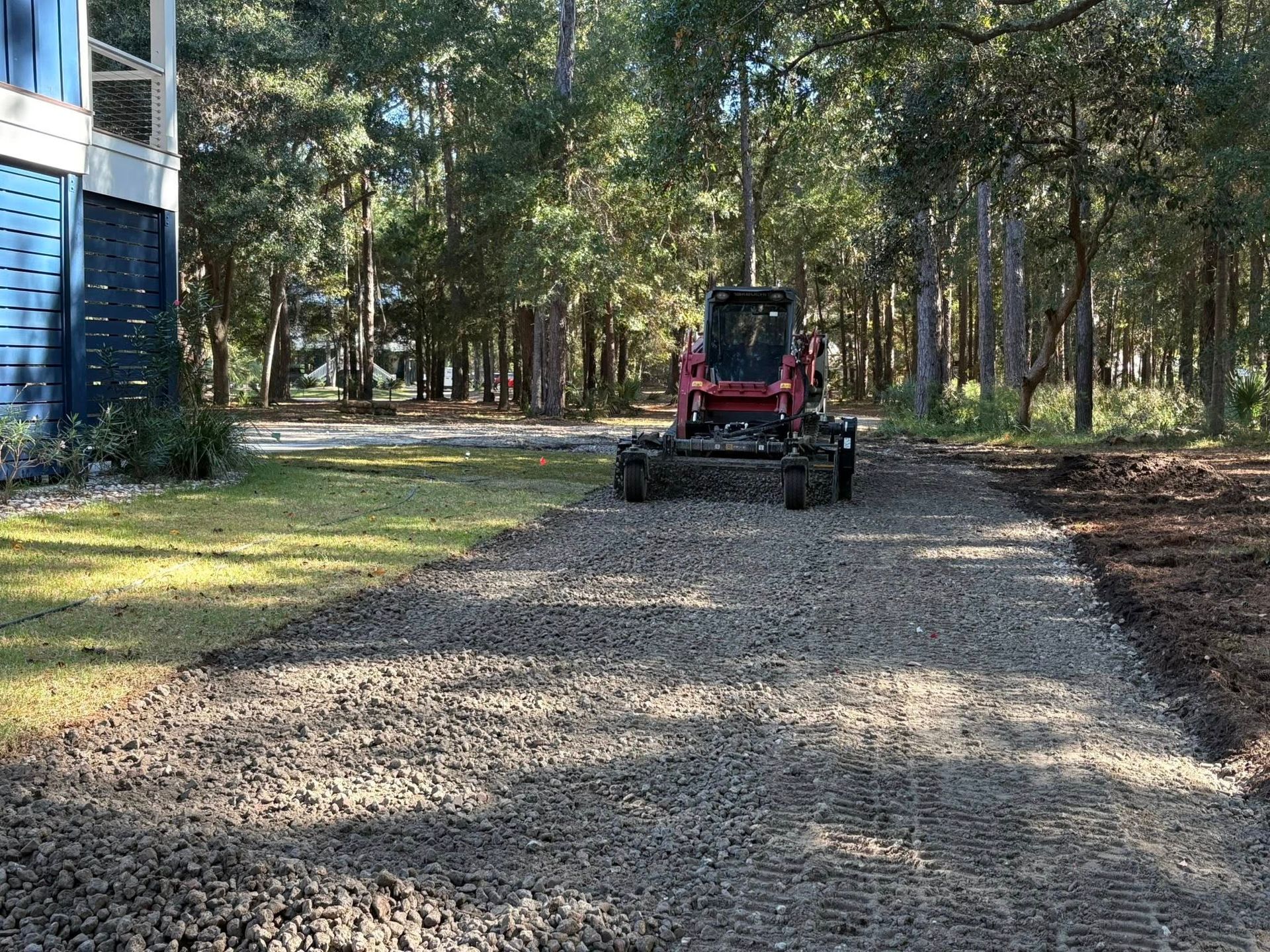 Red tractor on a gravel driveway beside trees and a house in a wooded yard