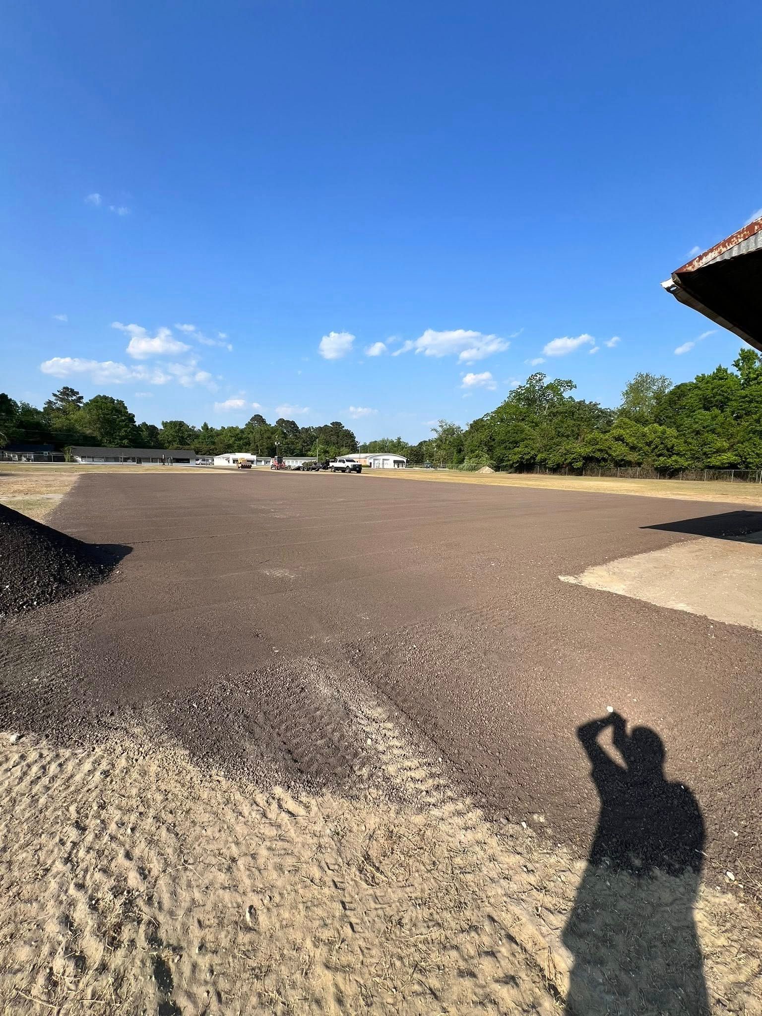 Gravel yard under a bright blue sky, with trees in the distance and a person’s shadow in the foreground
