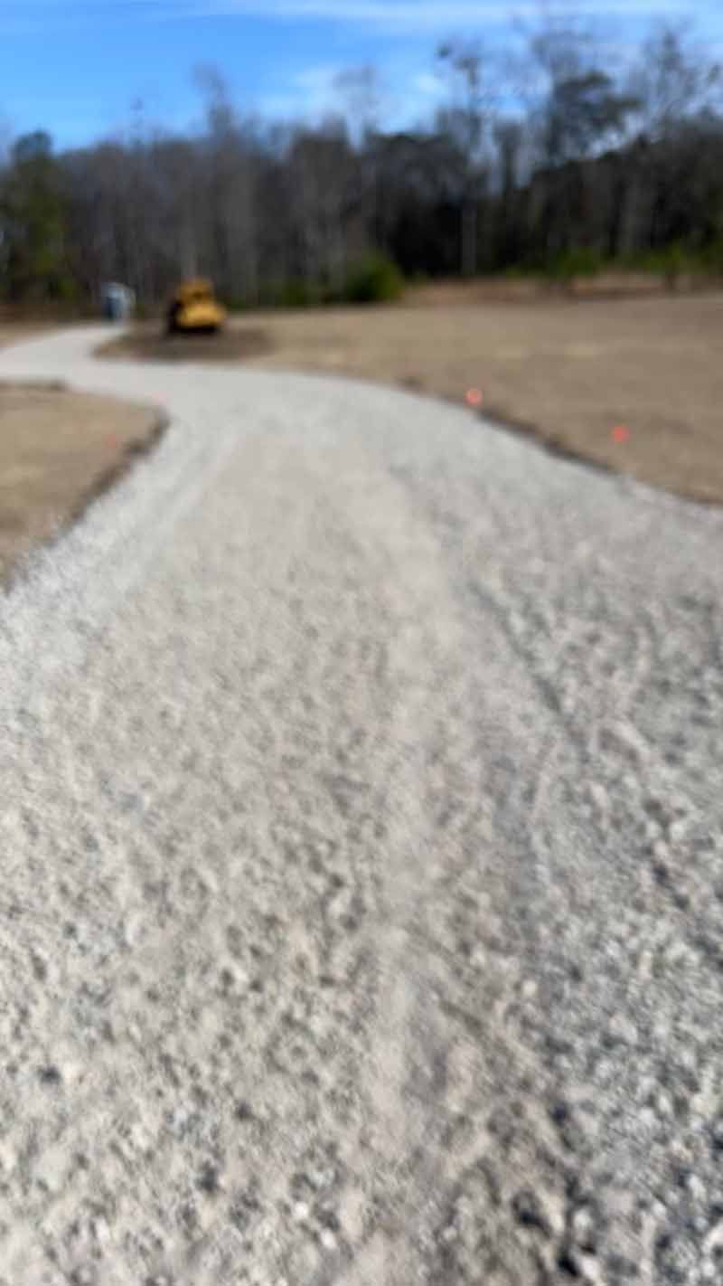 Gravel path curving through a cleared field with a small construction vehicle in the distance under a blue sky