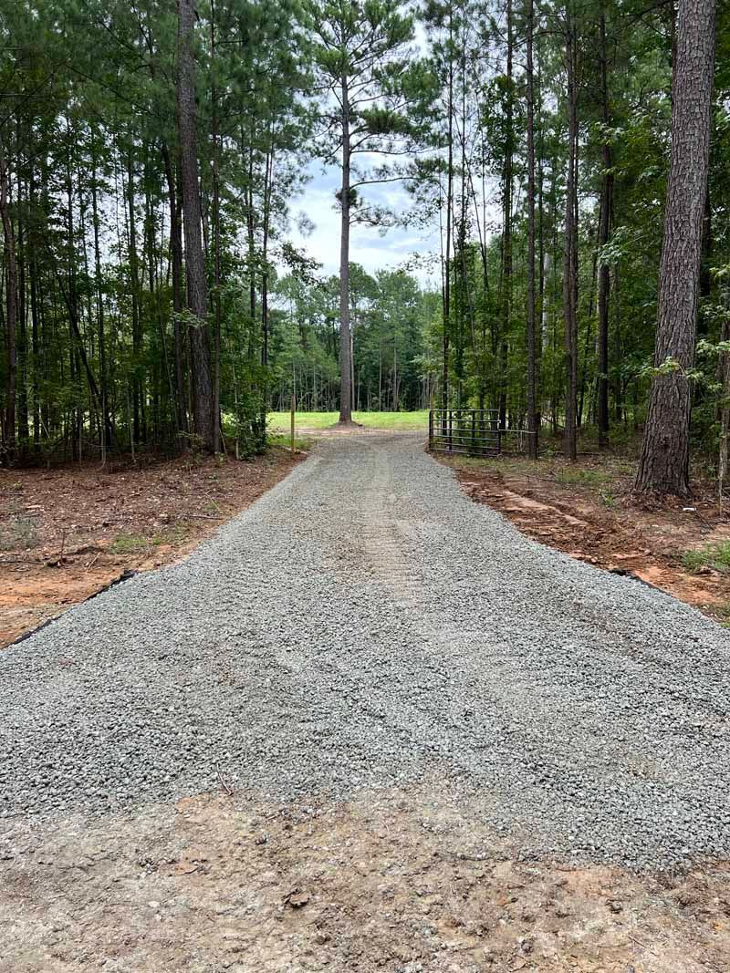 Gravel path through a wooded area leading to an open field under a cloudy sky