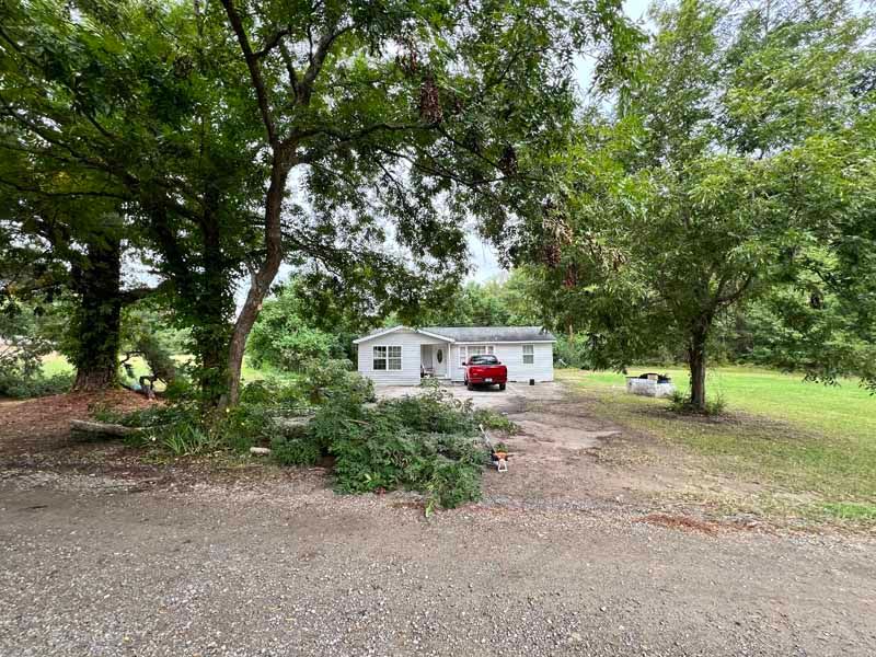 Small white house with a red car in the driveway, surrounded by large trees and a gravel yard.