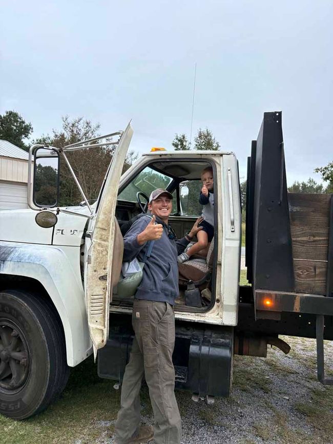 Man standing beside a white truck with open door and black dump bed, giving a thumbs-up outdoors.