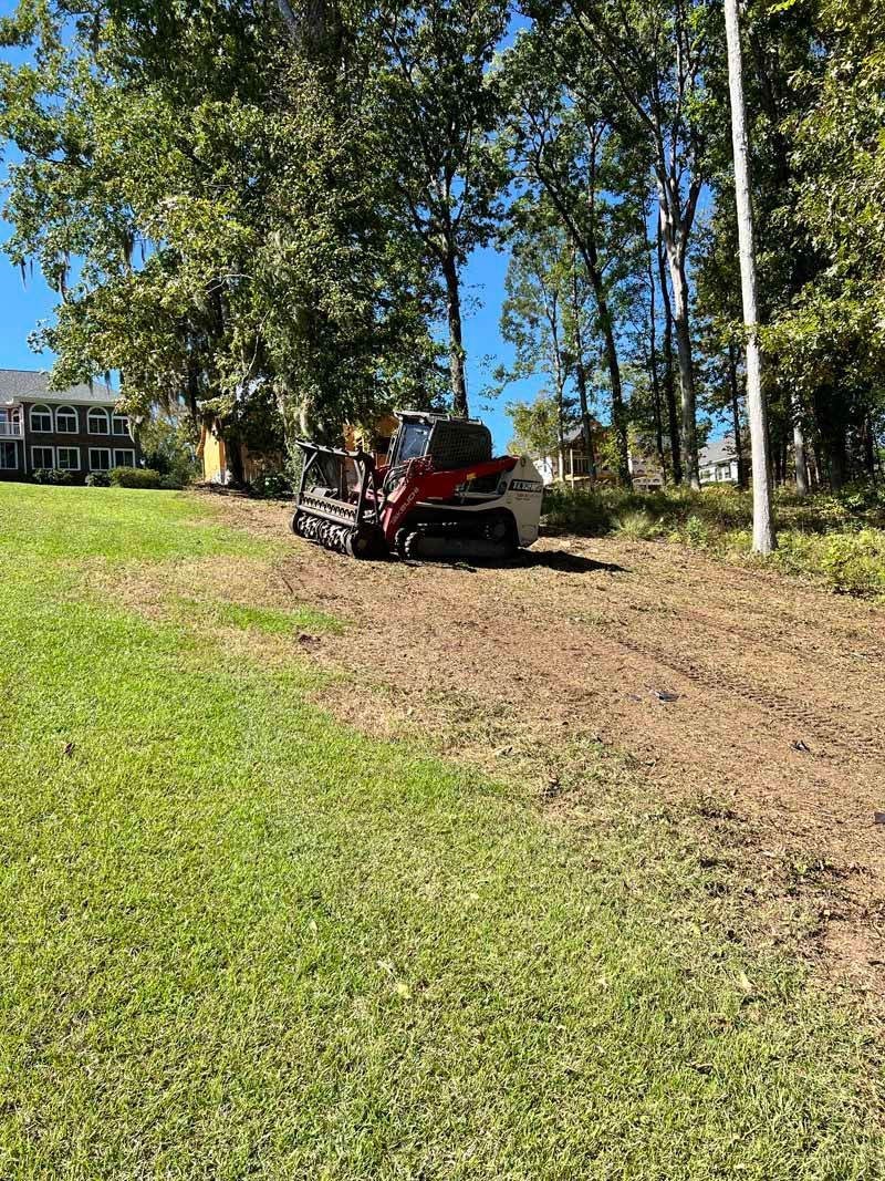 Red lawn mower clearing a dirt path beside a grassy yard with trees and blue sky