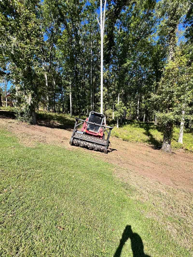 Brush cutter clearing a grassy slope beside trees and a fence line.