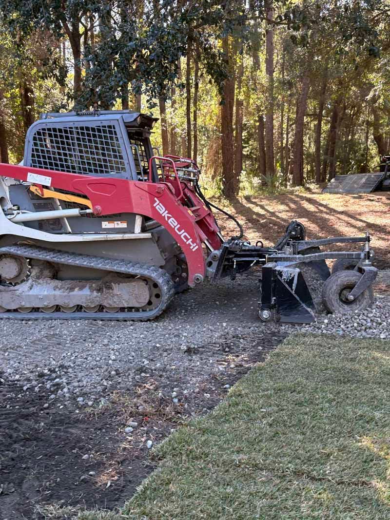 Red skid steer clearing gravel beside a wooded driveway under tall trees.