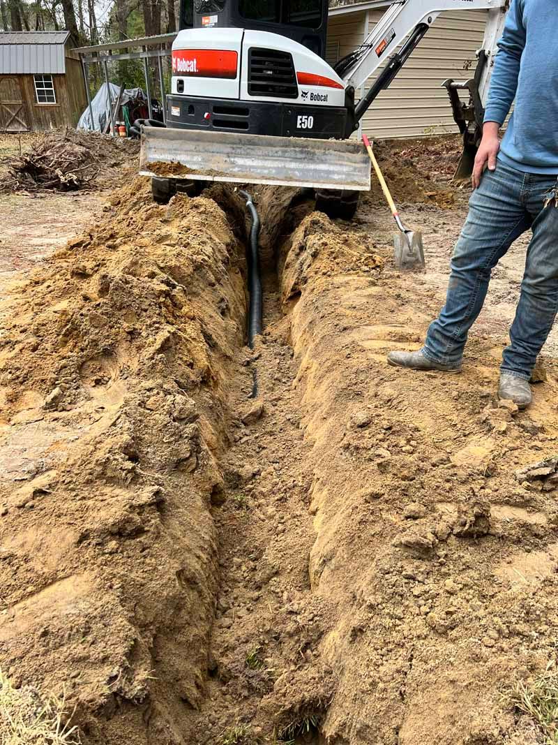 Excavator digging a deep trench in muddy ground beside a worker in jeans and boots.