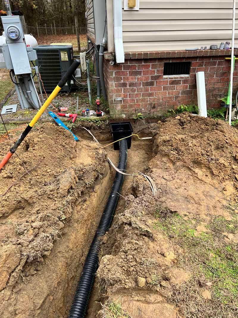 Trench with black corrugated pipe beside a house foundation and utility meters in a dirt yard
