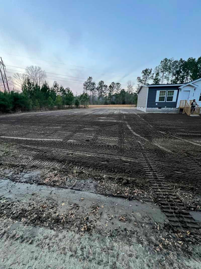 Freshly graded dirt lot beside a small house at dusk, with trees lining the background