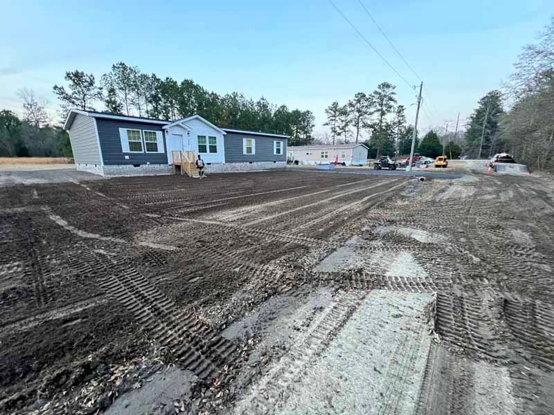 Newly graded dirt lot beside a gray manufactured home under a clear blue sky