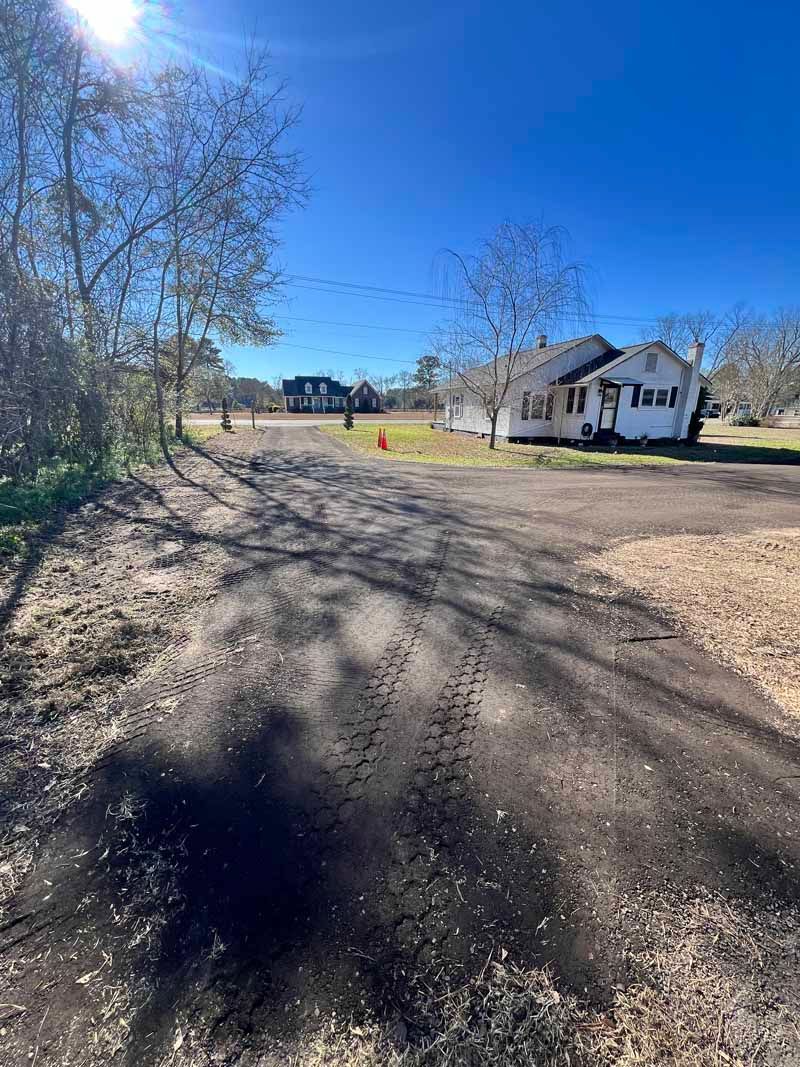 Dirt road beside leafless trees and a white house under a bright blue sky.