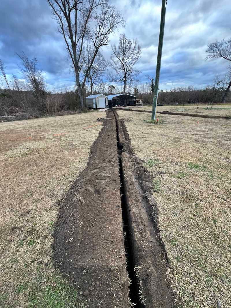 Long narrow trench cut through a grassy yard under a cloudy sky, with trees and utility poles nearby