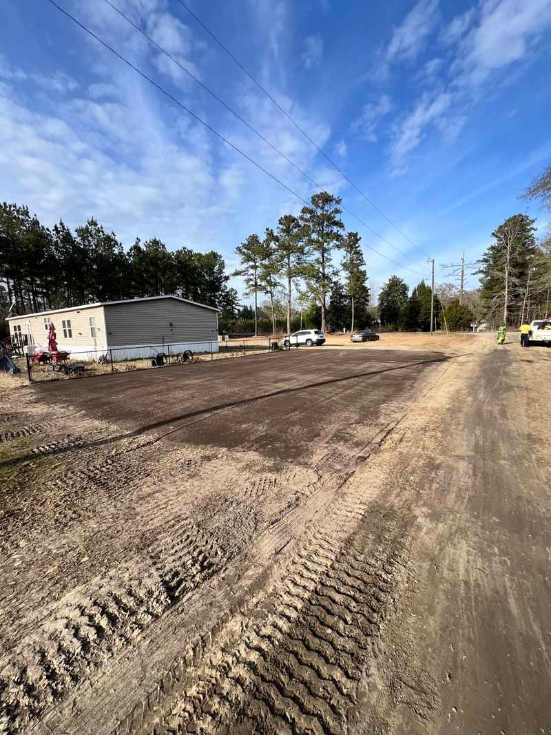 Dirt road construction site with tire tracks, a small white building, trees, and blue sky.