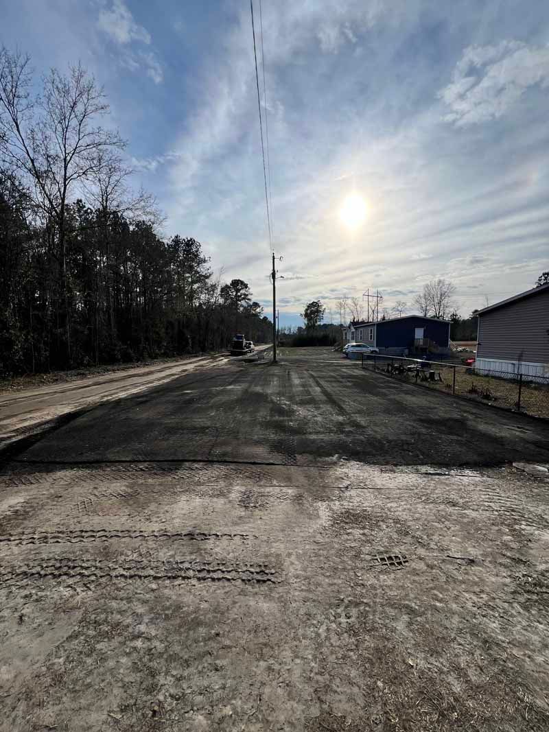 Sunlit gravel road beside trees and buildings under a hazy sky