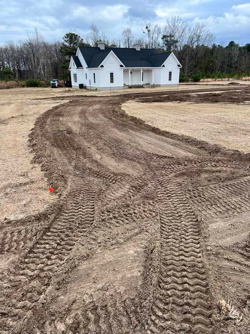 Tire tracks in muddy ground leading toward a small white house under a cloudy sky.