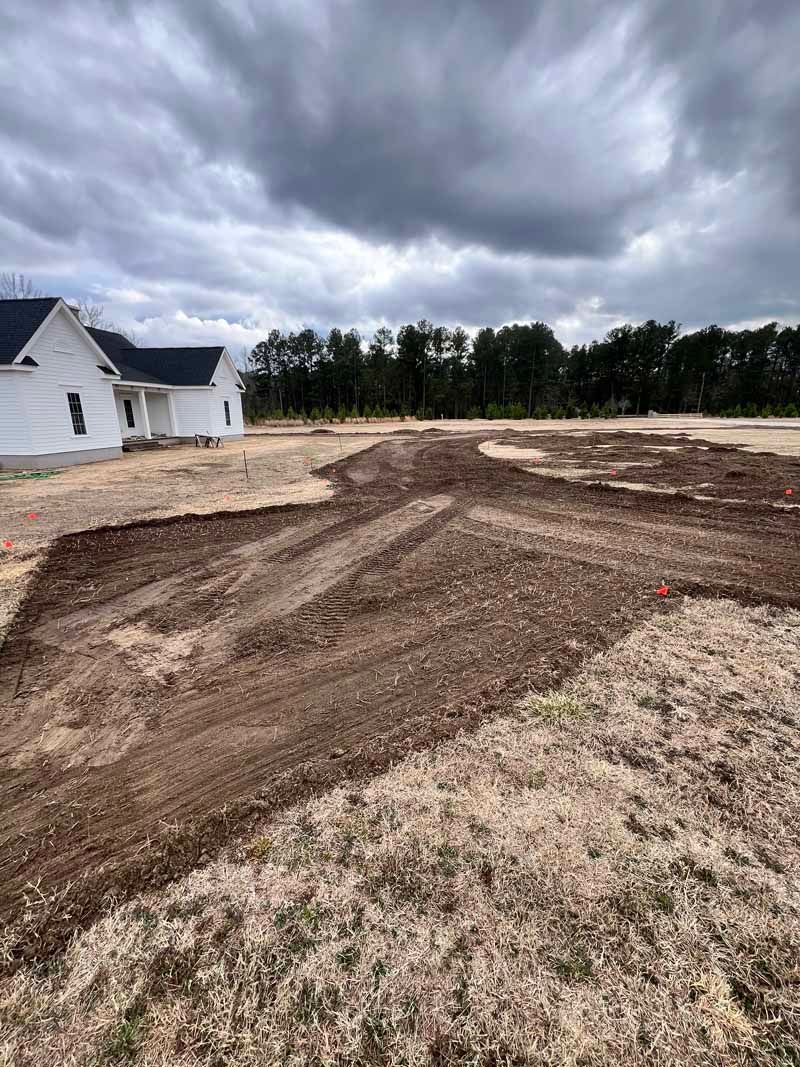Freshly graded dirt lot beside a white house under a cloudy sky