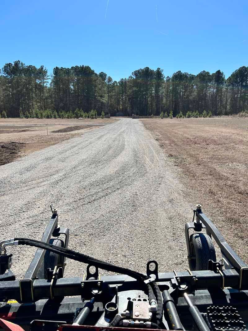 View from a vehicle on a gravel road through a dry field toward a tree line under a clear blue sky