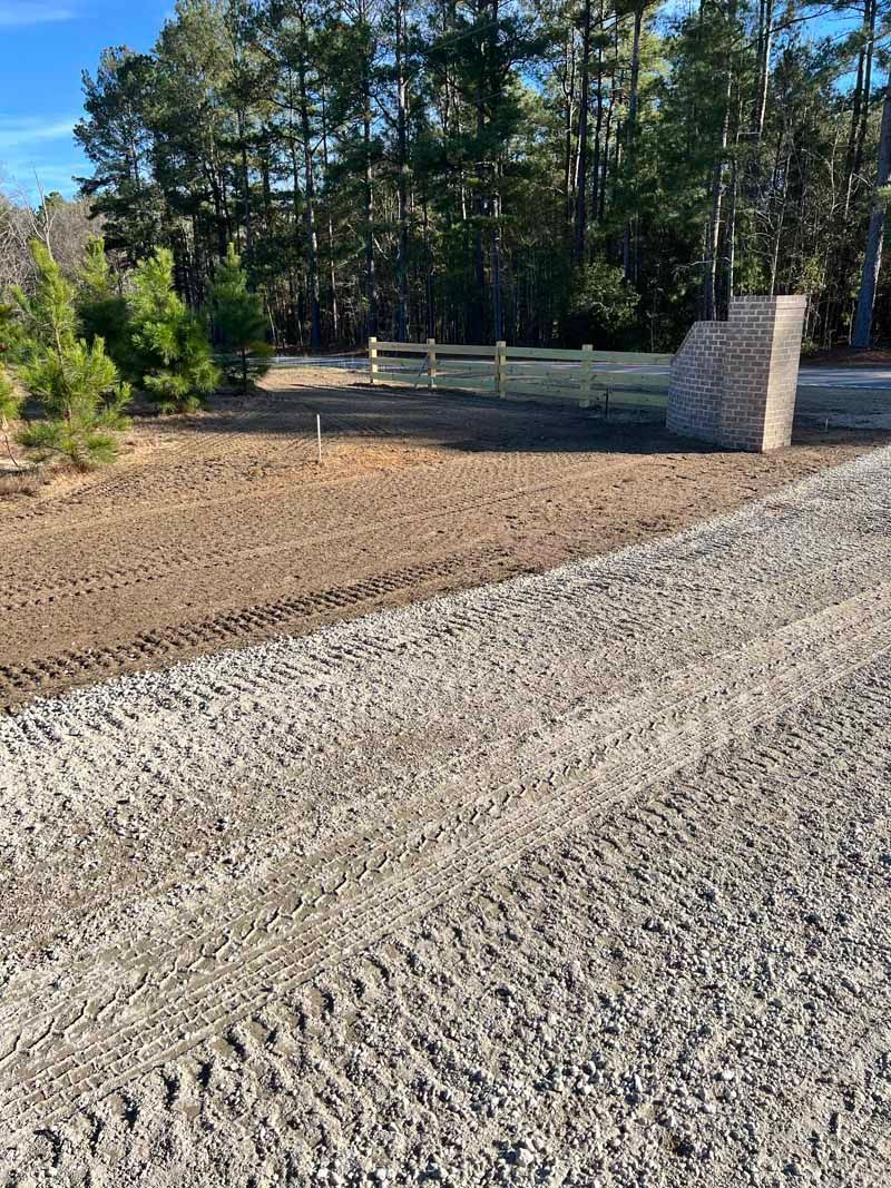 Gravel driveway leading to a landscaped lot with trees, dirt, and a stone pillar at the edge.