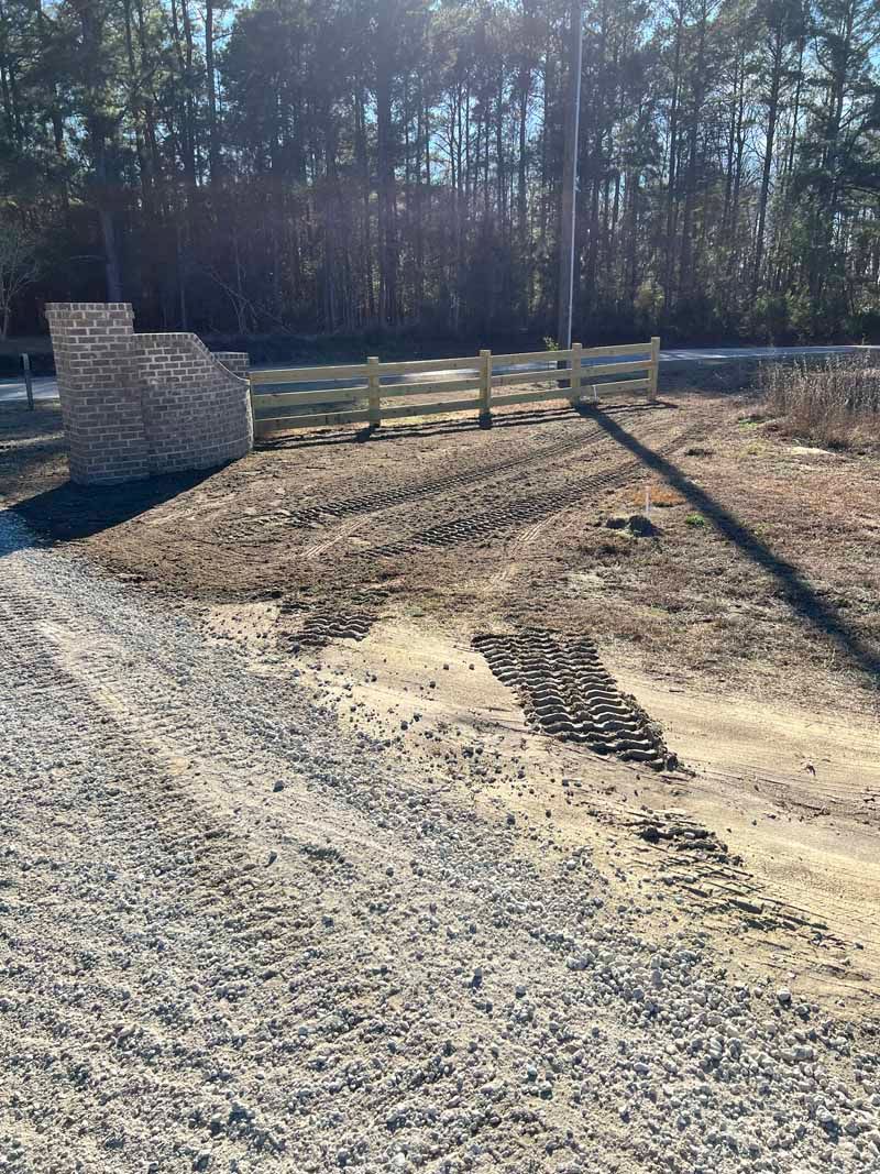Soil-covered lot with tire tracks, a low stone wall, and trees in the background