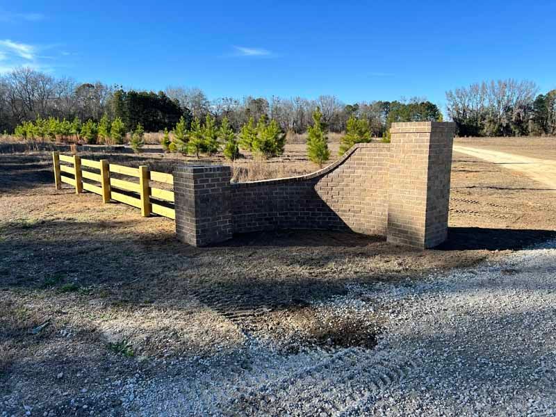Brick-and-wood entrance gate beside a gravel drive in a rural, tree-lined area