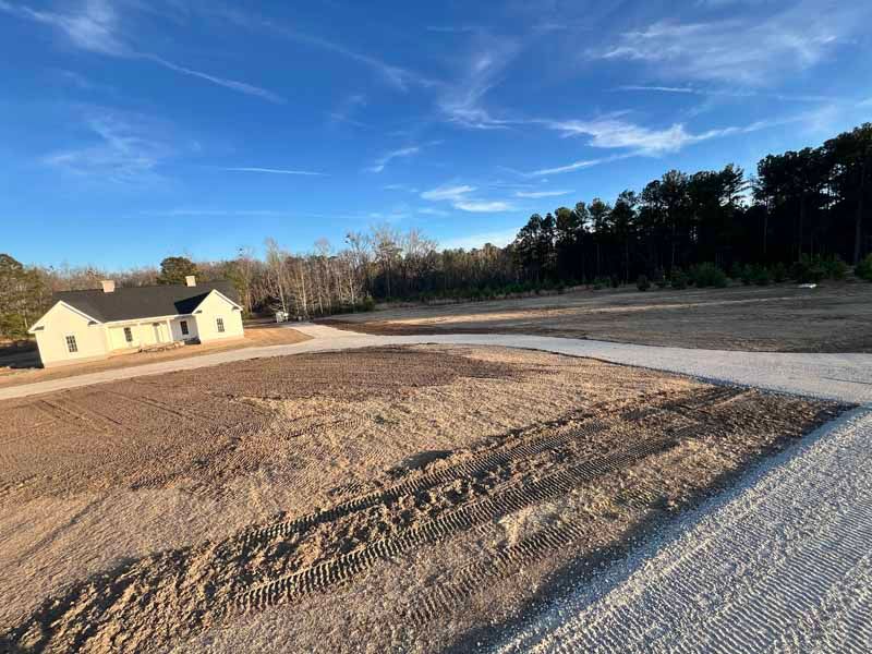 Gravel road leading to a small beige house on a cleared lot under a blue sky
