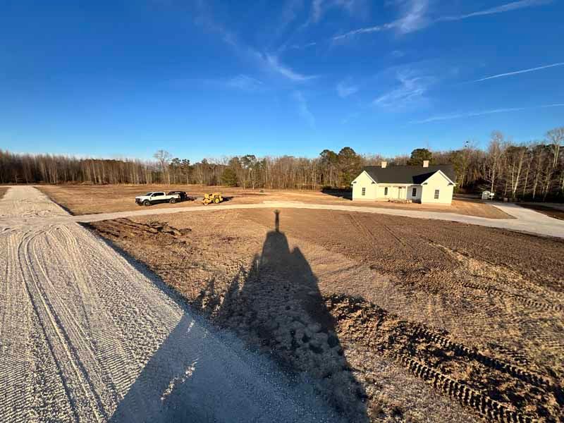 Wide dirt lot with a yellow house in the distance and a long shadow in the foreground under a blue sky