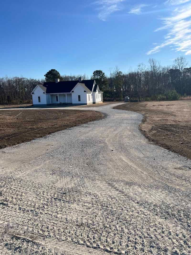 Gravel driveway leading to a white house with a dark roof in a wooded rural setting under a blue sky