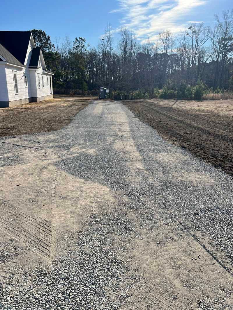 Cracked gravel driveway beside a white house, leading toward bare trees under a blue sky.