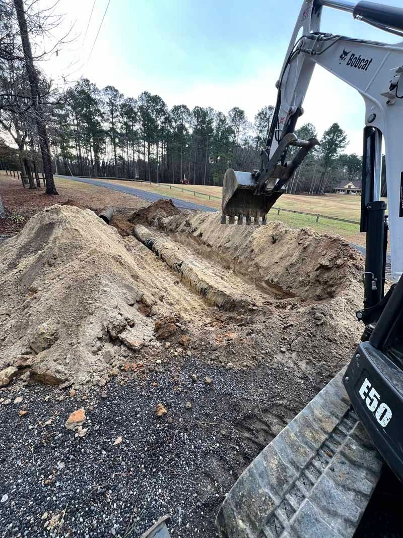 Excavator digging a dirt trench beside a wooded field during construction