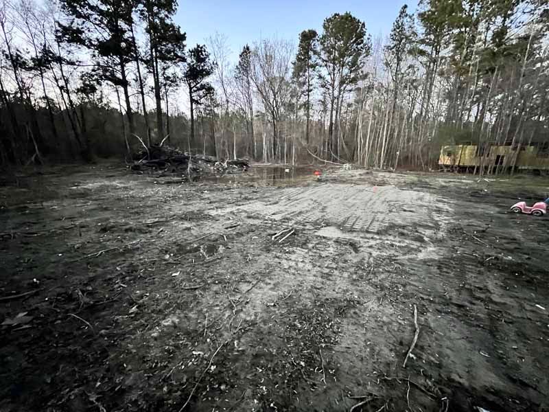 Cleared dirt lot in a wooded area with trees around the edges and a small yellow structure in the distance
