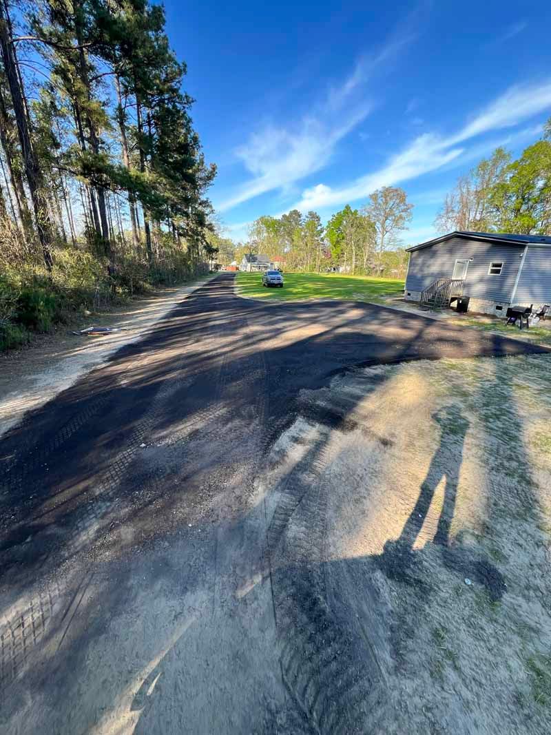 Dirt road by trees and a small building under a blue sky, with long shadows in the foreground