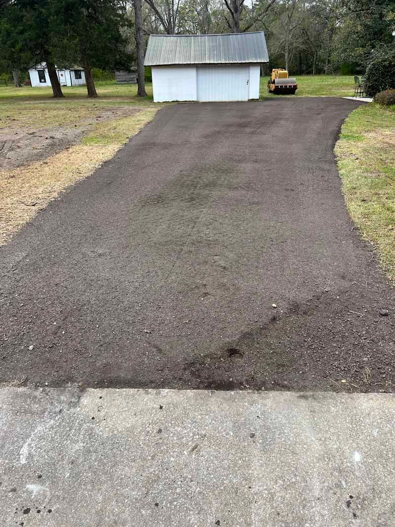 Gravel driveway leading to a small white shed in a wooded yard, with a concrete edge in the foreground.